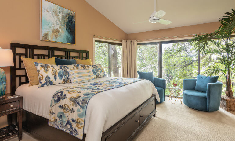 View of bedroom look toward corner with windows in L-shape surrounded by woods, cherry furniture, blue chairs by windows and palm tree in wicker pot.