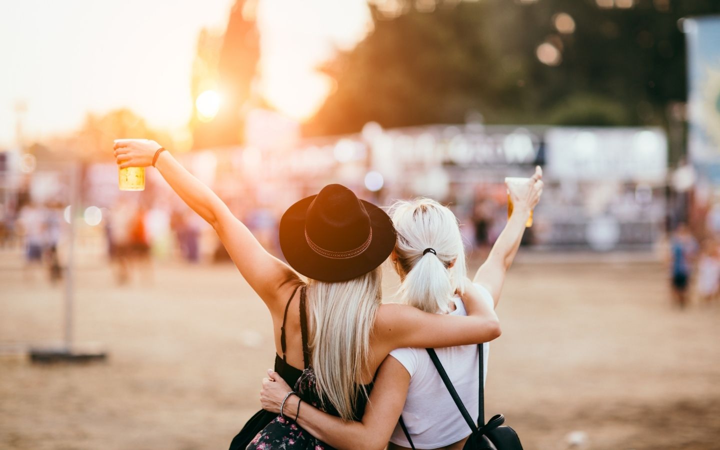 Back side of two women, one with a cowboy hat, raising their beer cups toward live music